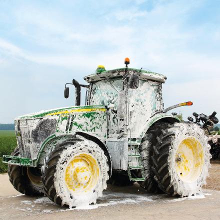 A green and yellow tractor covered in white soap suds is being washed outdoors on a concrete surface near a field.