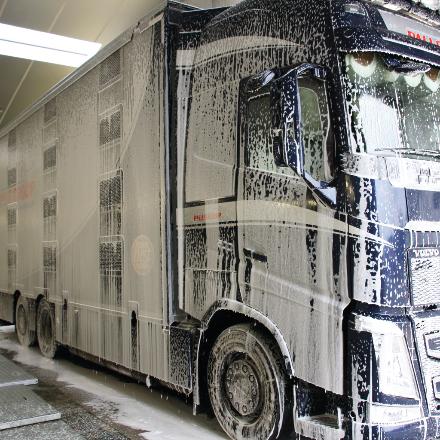 A large black Volvo truck is covered in white soap suds while being washed inside a garage or washing facility.