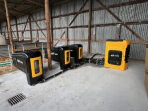 Three black animal feeding stations are connected by cables to a large yellow control unit inside a metal-roofed farm building.